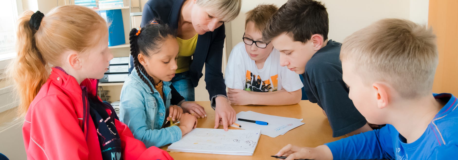 Fünf Schüler hören den Erklärungen der Lehrerin aufmeksam zu. Arbeitsblätter liegen auf dem Tisch.