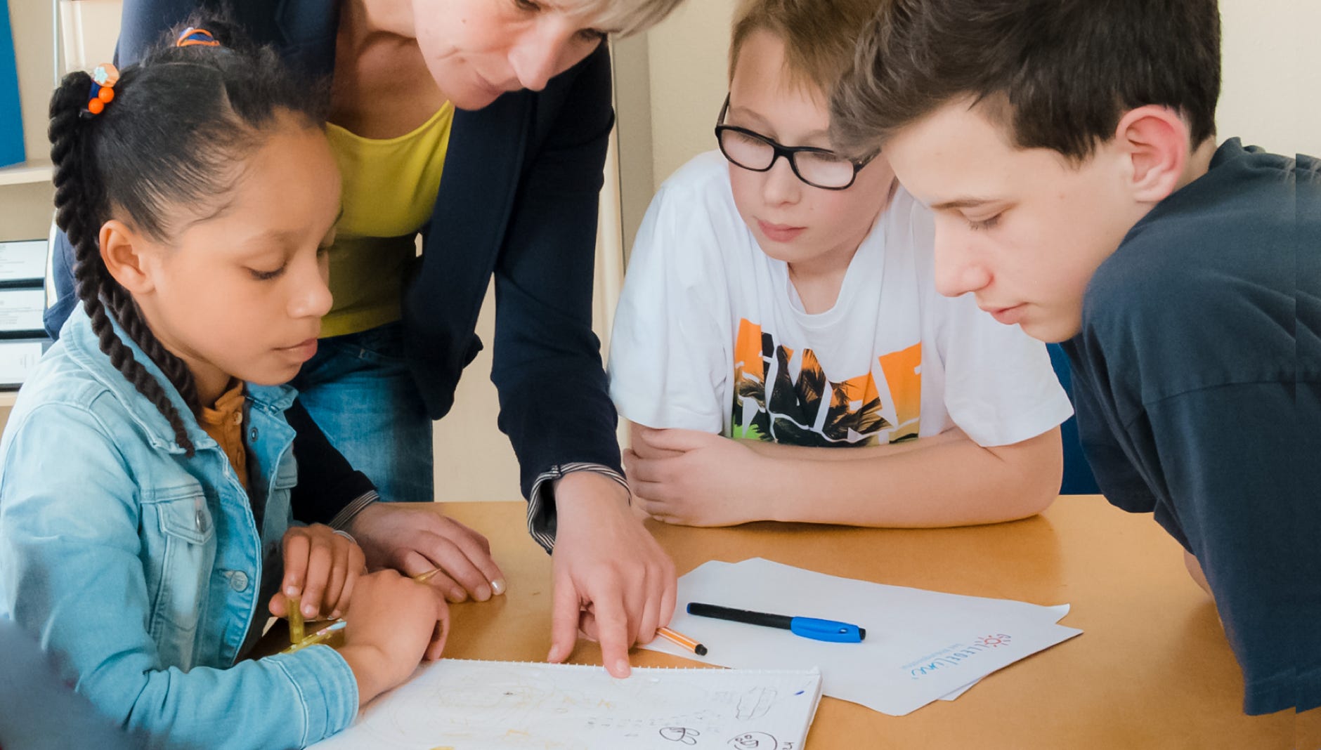 Lehrerin erklärt 3 Schülern eine Aufgabe. Die Kinder hören interessiert zu.